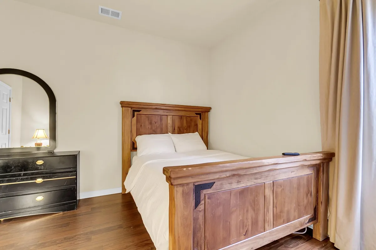 A peaceful bedroom featuring a wooden bed frame with fresh linens, a black dresser, decorative mirror, and curtains softly framing the window. The room is illuminated by natural light, creating a warm and tranquil atmosphere.