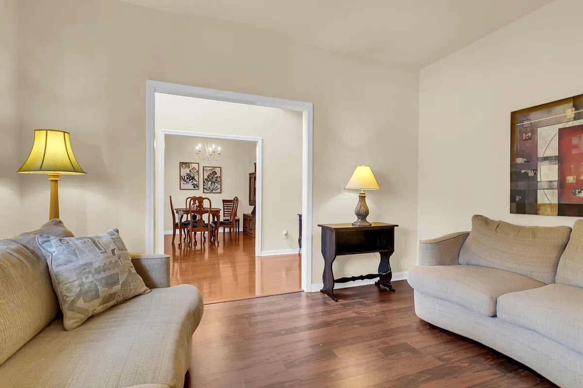 A bright, comfortable living space with a sofa, table lamp, a glimpse into the dining room, and wall art, ideal for socializing, reading, or enjoying gatherings.