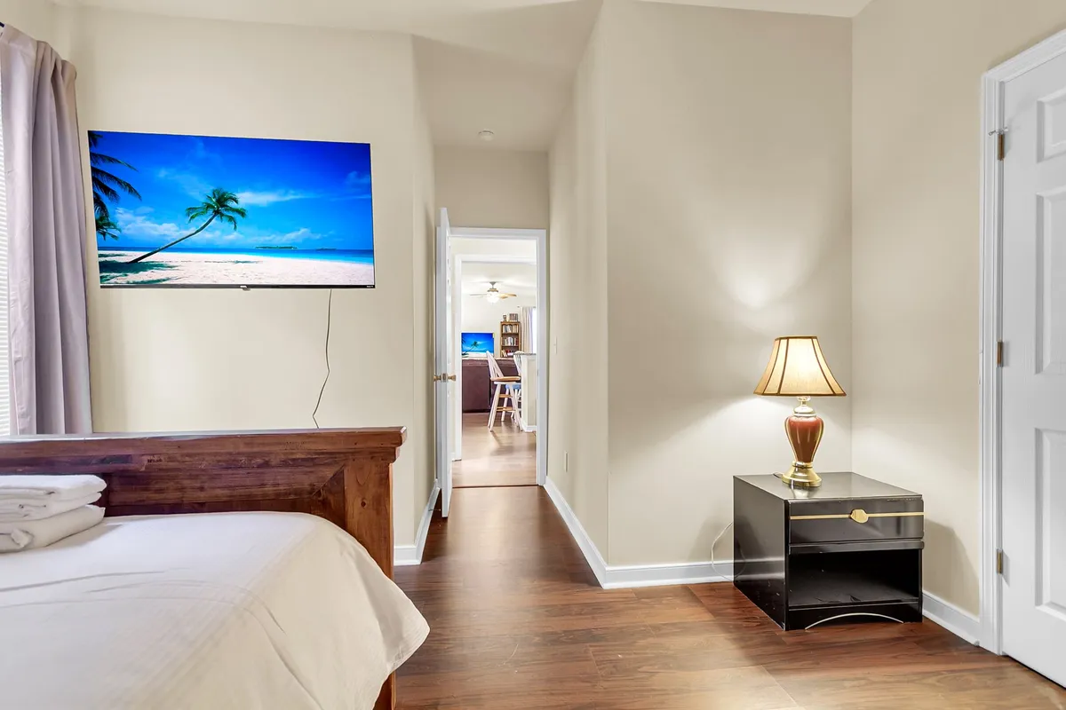 A cozy bedroom with a bed neatly arranged with fresh linens, a TV displaying a soothing beach scene, a lamp on the nightstand, and an open doorway hinting at spaciousness. The ambiance is relaxed and inviting, ideal for unwinding.
