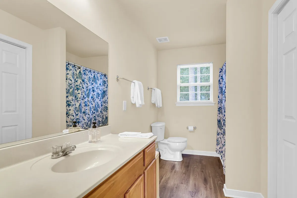 A calming bathroom scene featuring a vanity with sink, toilet, window streaming in daylight, and a floral shower curtain. The ambience is refreshing and serene, ideal for self-care and private routines.
