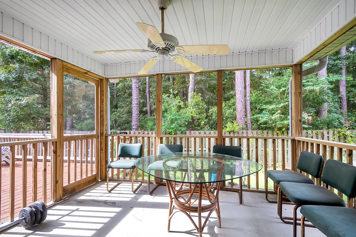 The image showcases a tranquil, sun-dappled dining area featuring a glass table surrounded by inviting green chairs and warm wooden railings. Gentle morning light filters through the trees outside, while a ceiling fan and open layout hint at both comfort and relaxation, evoking the feeling of a peaceful family retreat nestled in nature.