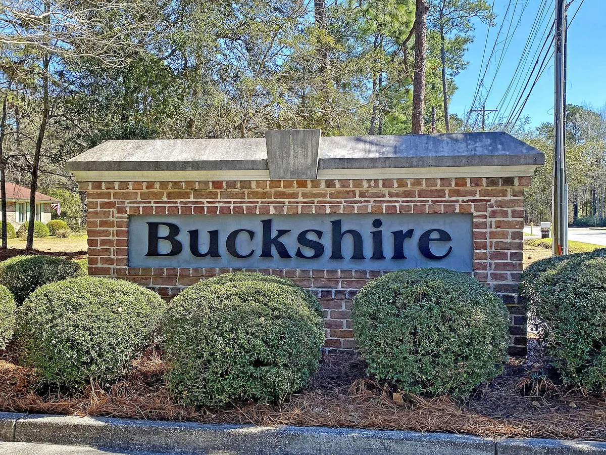 The Buckshire neighborhood sign surrounded by landscaped shrubs and mature trees. The scene presents a welcoming entrance to the community, evoking a sense of belonging and safety for families exploring the area.